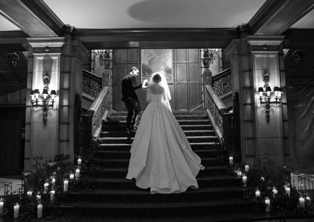 Black and white image of groom and bride walking up stairs in luxurious wedding venue.